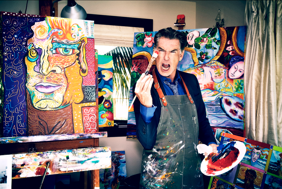 Pierce Brosnan in his art studio wearing an apron and holding a paintbrush and palette, surrounded by his colorful expressionist paintings, capturing his passion for visual art.