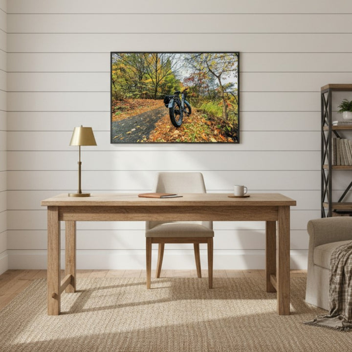 Home office with wooden desk, chair, lamp, and framed picture of a fat bike on a forest path.